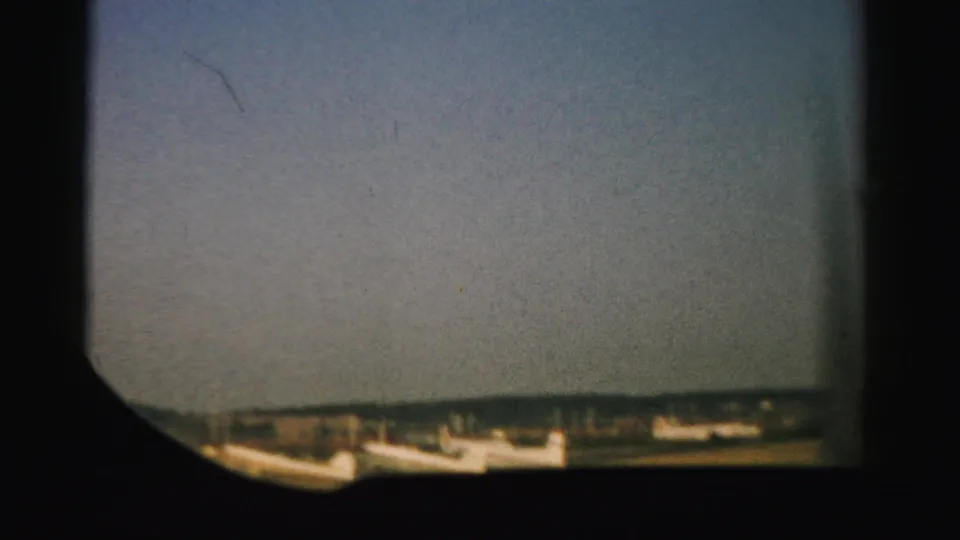 From an aircraft window, the 1957 Nevada footage looks across runway ground and small distant airport structures.