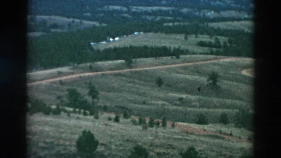 1958 8mm frame from an elevated moving viewpoint over rolling green fields, pale dirt roads, scattered trees, and distant structures.