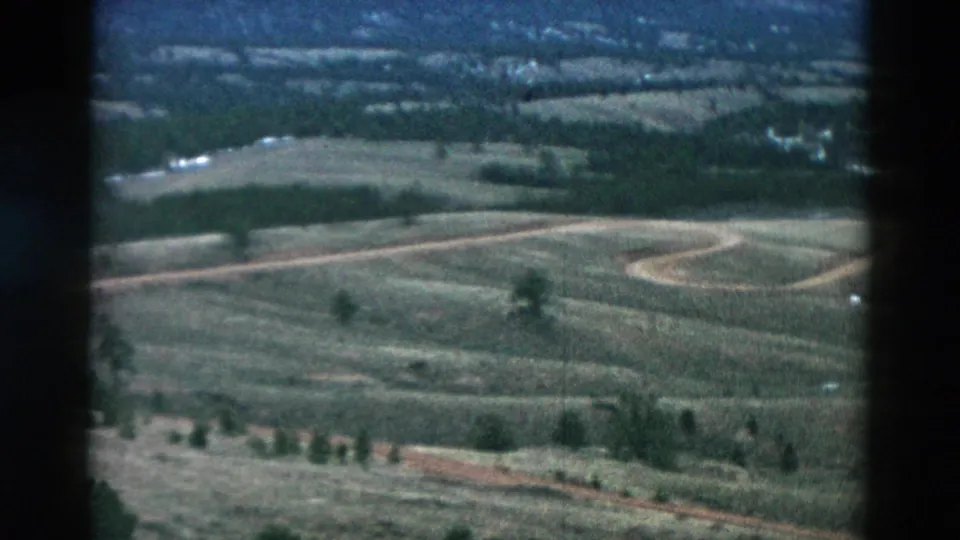 1958 8mm view across rural fields with a looping dirt road, tree rows, pale ground patches, and soft distant landscape detail.