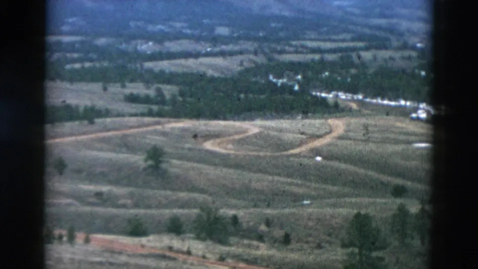 Wide 1958 8mm frame showing an elevated rural vista with fields, winding dirt road, scattered trees, and small distant structures.