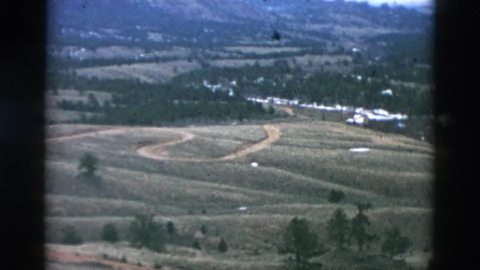 1958 archival 8mm aerial view of a rural valley, with open fields, curved dirt roads, sparse trees, and small distant buildings.