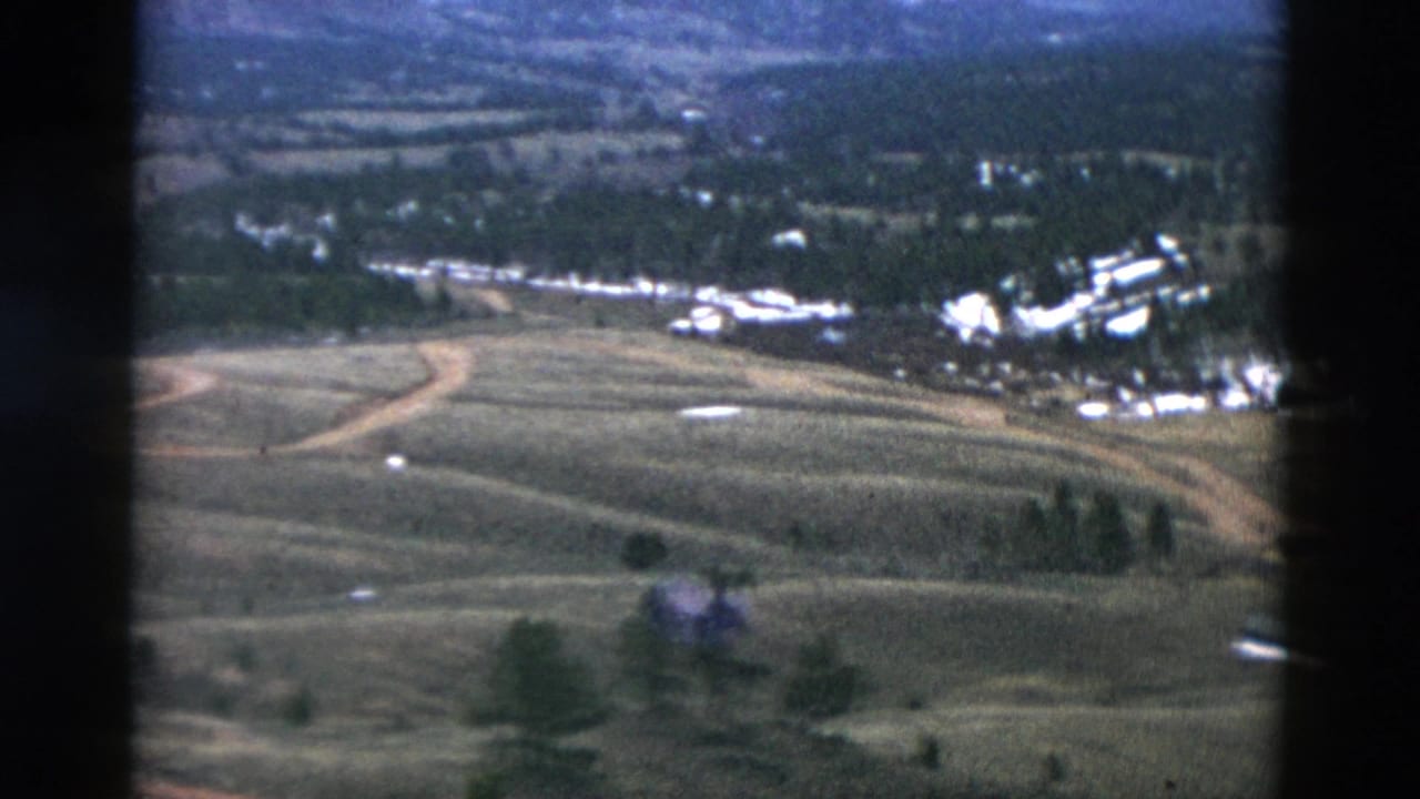 Aerial frame of rolling open land, curved roads, tree patches, and pale buildings in the distance.