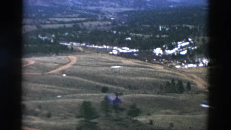 Archival 8mm aerial footage over rural Oregon open land, with rolling fields, a curving dirt road, trees, and distant buildings.