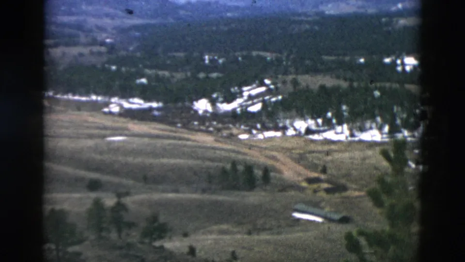 Mid-century 8mm aerial view of open rural land, with a road junction, scattered trees, and light-colored buildings beyond the fields.