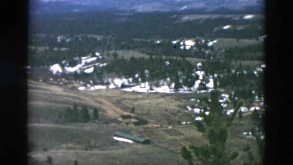 1958 archival 8mm aerial frame showing rural open land, a curving road, trees, distant buildings, and a pale elongated foreground shape.