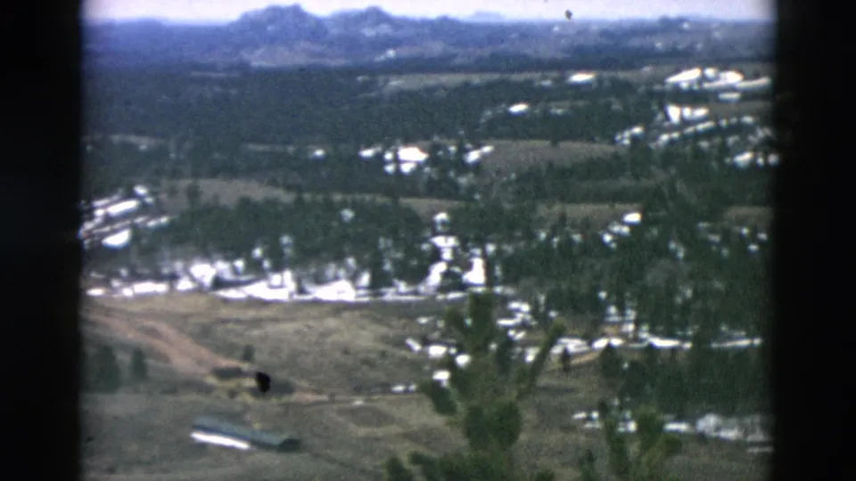 1958 8mm aerial view described as Oregon, showing snow-dappled woods, open fields, buildings, and low mountains in the distance.