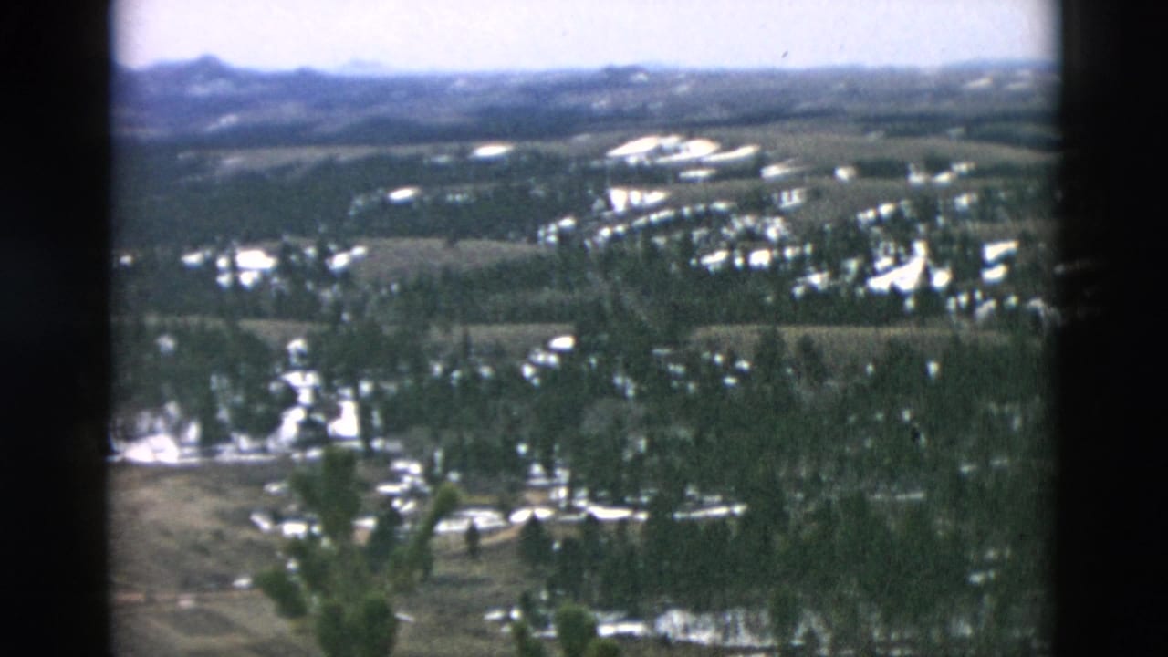 Aerial landscape with forest, snow patches, fields, buildings, and distant ridgelines.