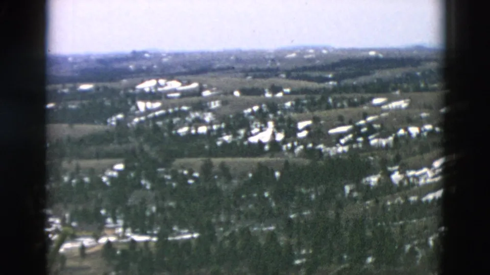 Soft 8mm aerial frame showing a broad rural landscape with woods, snow traces, buildings, open ground, and distant hills.