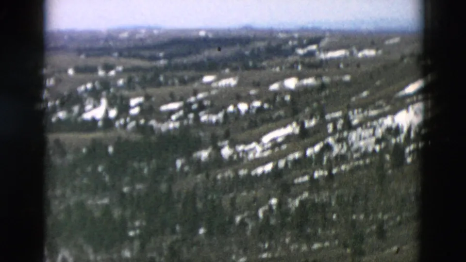 End frame of an 8mm aerial sweep across snow-dappled woods, rural structures, open land, and low hills under a pale sky.