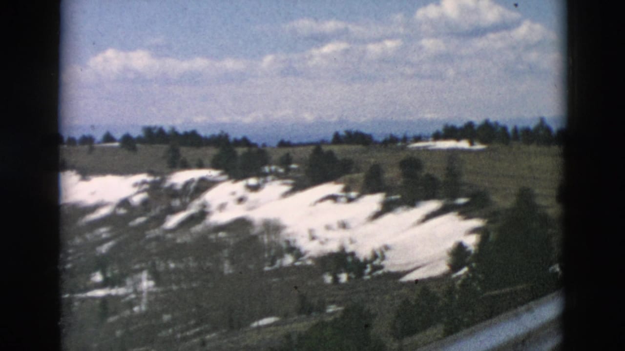 Snow-patched hills with scattered trees under a blue sky and clouds.