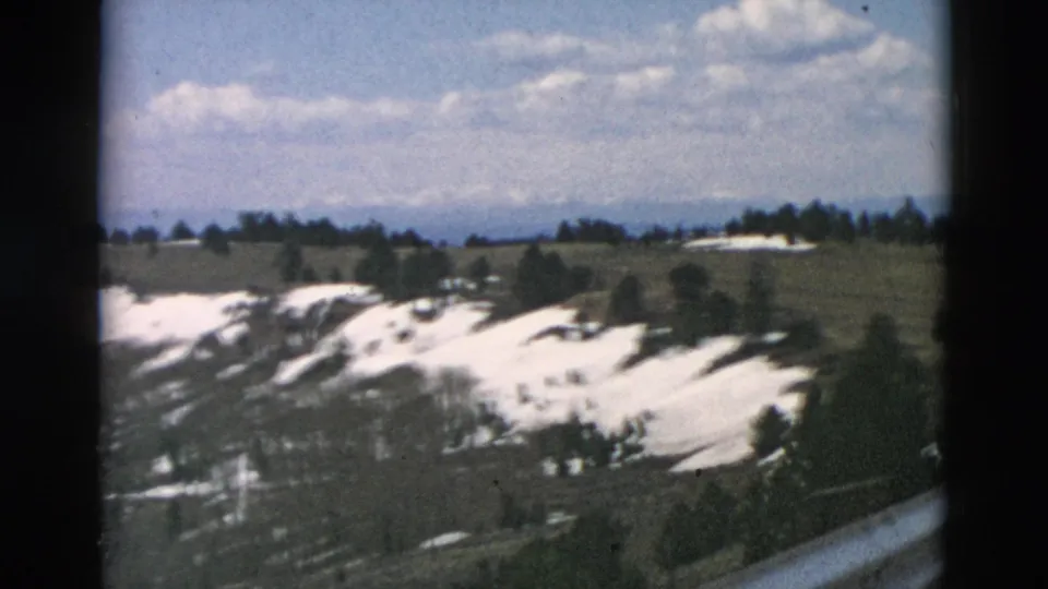 1958 8mm frame of open mountain hills with lingering snow, sparse trees, distant ridges, and scattered clouds.