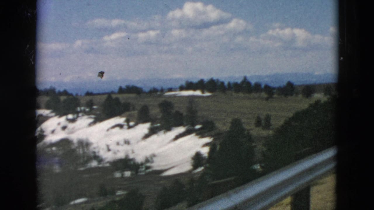 Roadside 8mm view of snow-patched hills, sparse conifers, distant ridges, and a guardrail in the foreground.