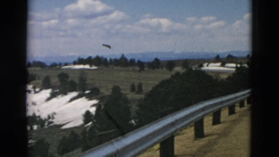 Home-movie frame looking across snow-streaked mountain fields with trees, distant horizon, clouds, and roadside railing.