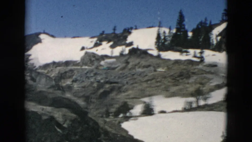 8mm view of exposed rock, lingering snow, and conifers on a mountain slope beneath a clear blue sky.