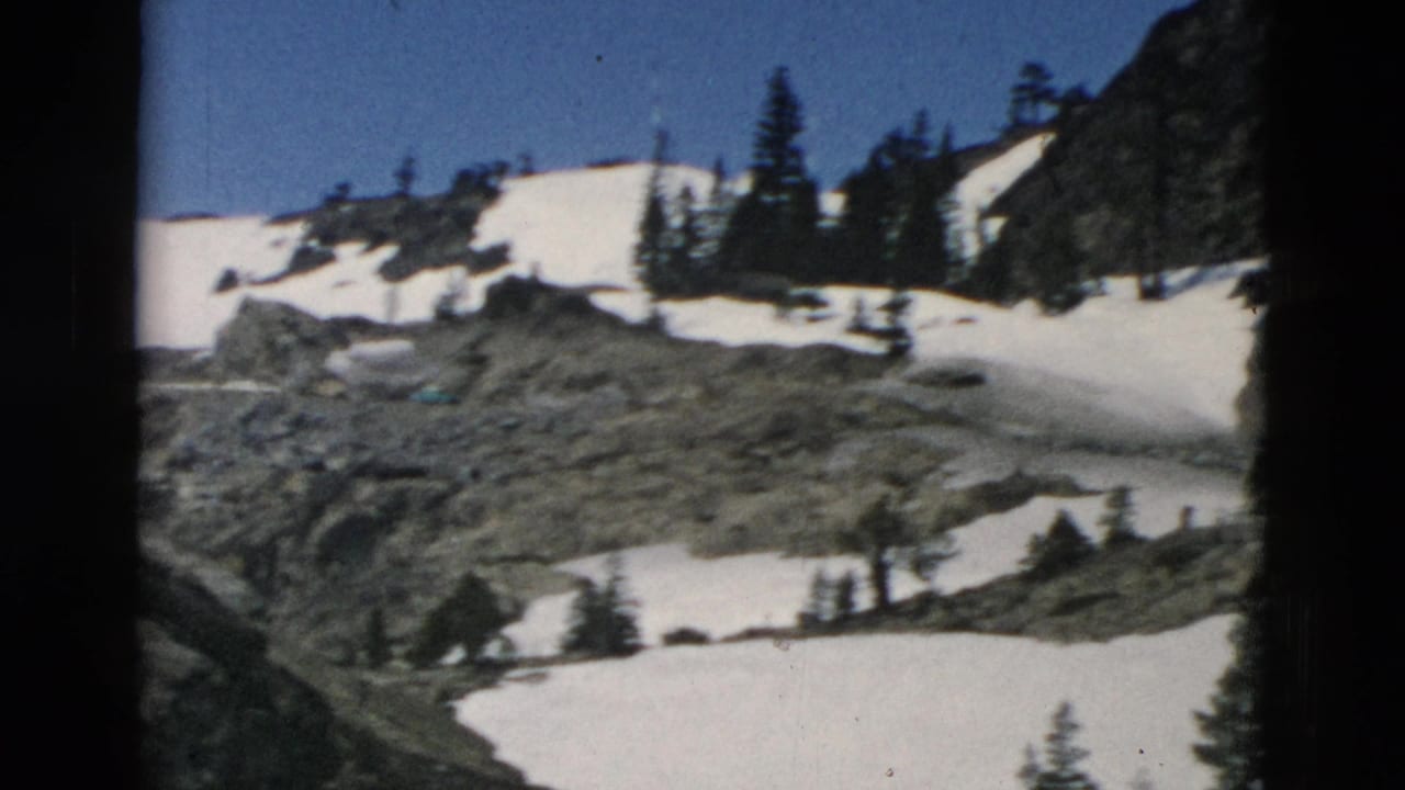 Snow patches on rocky mountain slopes with evergreen trees under a blue sky.