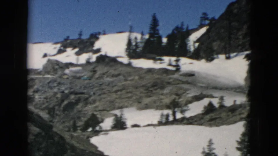 Archival 8mm frame showing rocky mountain terrain, lingering snowfields, scattered evergreens, and a clear blue sky.