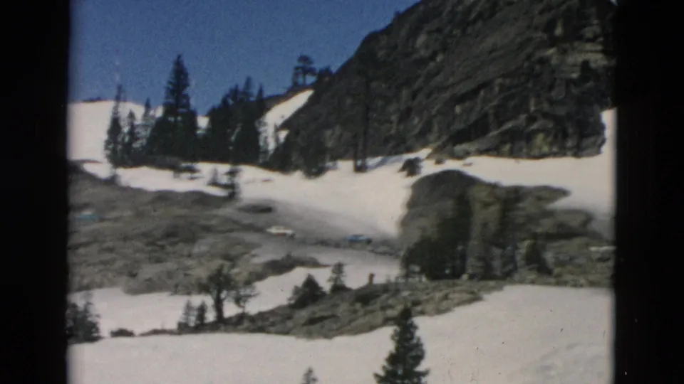 Archival 8mm frame with snowfields, rock outcrops, evergreen trees, and a mountain slope partially obscured by the film frame edge.