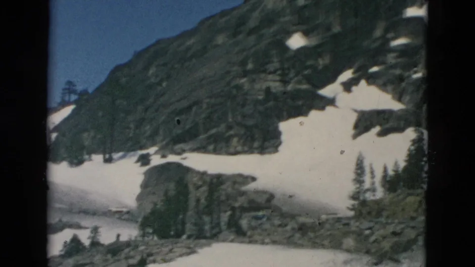 Archival 8mm frame showing a darker rocky mountainside, broad snow patches, and scattered evergreens during the pan.