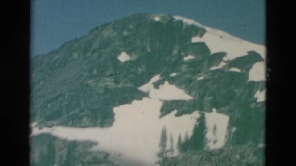 Archival 8mm frame with a green color shift, showing a snow-dappled mountain slope and dark evergreen trees near the bottom.