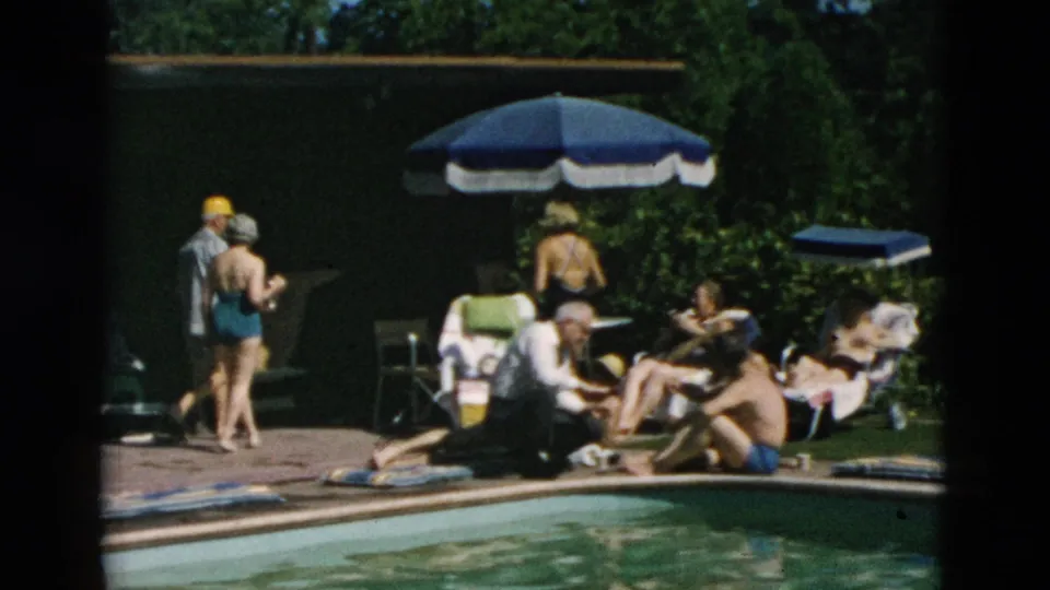 1958 home-movie frame showing adults around a pool, with lounge chairs, sun hats, and a blue-and-white umbrella nearby.