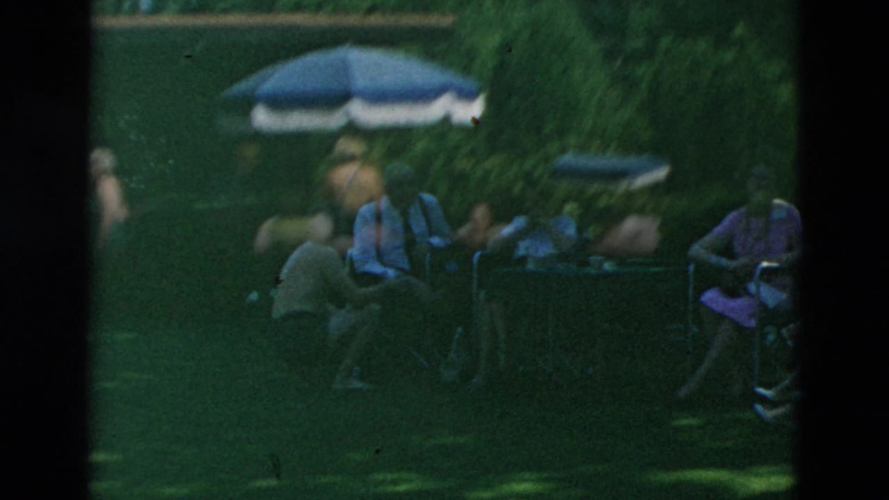 Shaded 8mm frame of adults seated on a lawn near a table, with a patio umbrella and dense greenery behind them.