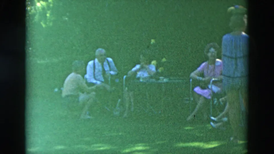 Adults sit and converse around a lawn table in a soft, green-tinted 8mm frame with trees and dappled light.
