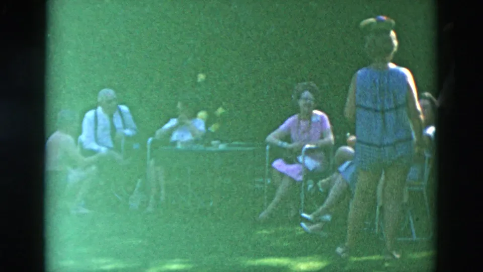 1958 home-movie frame showing adults in summer clothing gathered near a lawn table, with one person standing in the foreground.