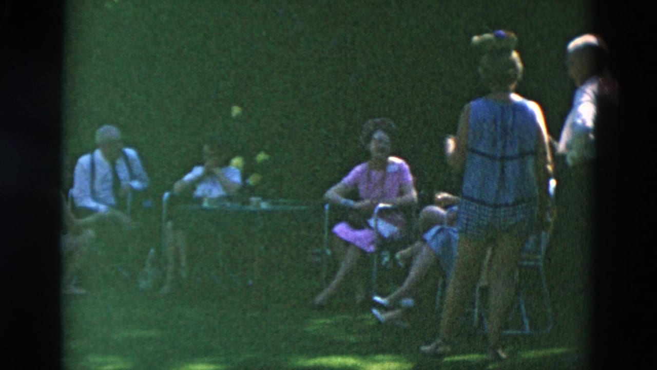 1958 Oregon home-movie frame showing adults in period clothing seated around a small table while others stand nearby on the lawn.