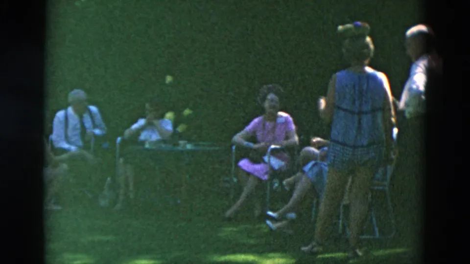 1958 Oregon home-movie frame showing adults in period clothing seated around a small table while others stand nearby on the lawn.