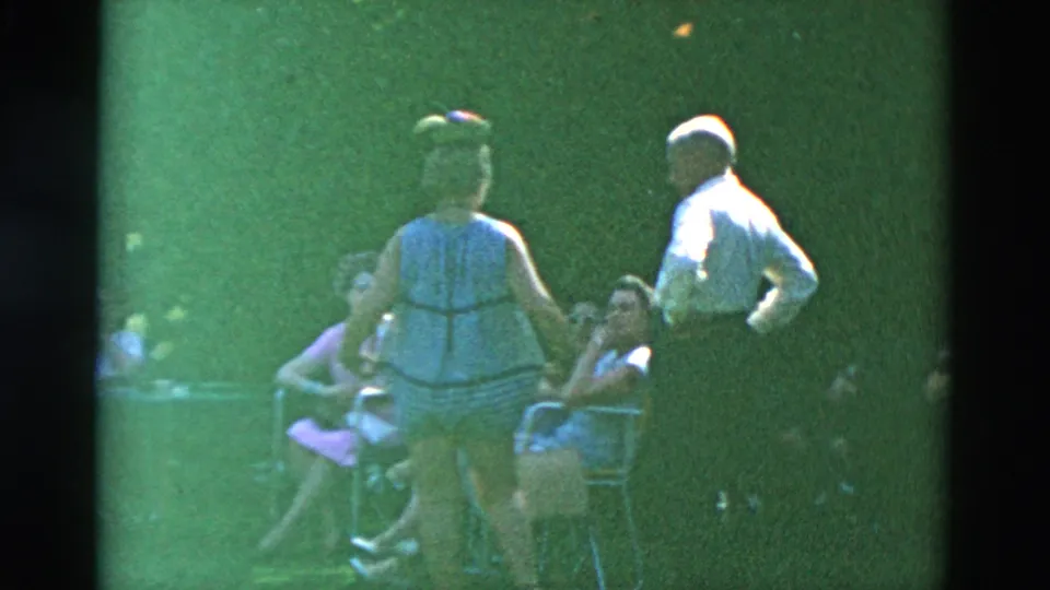 1958 Oregon home-movie frame of adults socializing outdoors, with two standing figures partly blocking seated guests in lawn chairs.