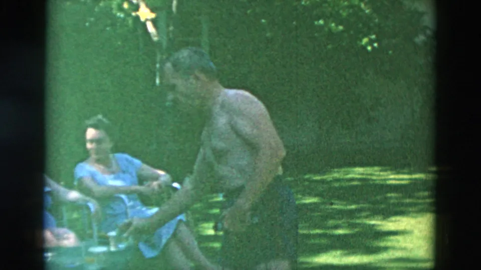 1958 Oregon backyard frame showing a man moving through the foreground while women sit in folding chairs on the grass behind him.
