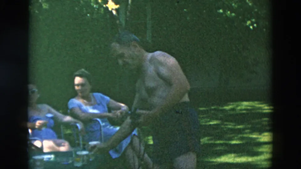 Archival 8mm frame of adults gathered outdoors in shade, with a shirtless man standing near seated women in blue clothing.