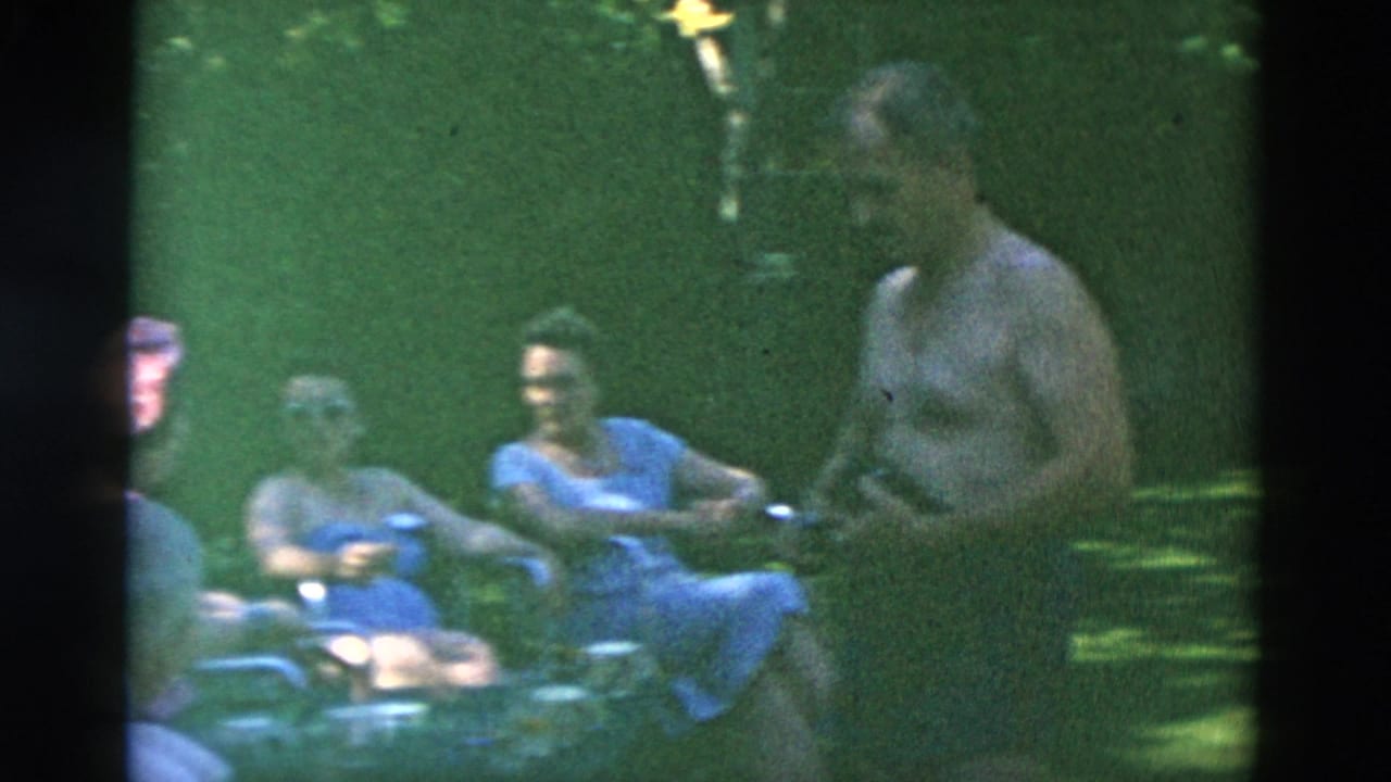 Adults appear gathered in a shaded outdoor area, with a shirtless man beside women seated in lawn chairs.