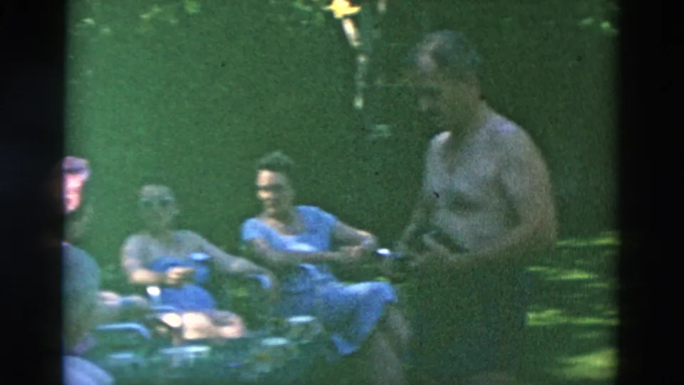 Adults appear gathered in a shaded outdoor area, with a shirtless man beside women seated in lawn chairs.