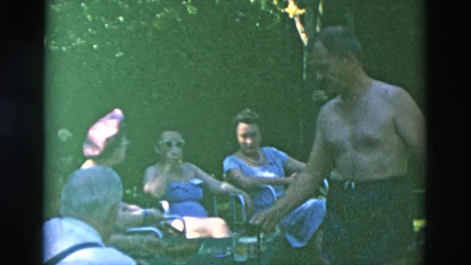 Shaded home-movie frame showing adults seated in lawn chairs, one in sunglasses and another with a pink patterned head covering.
