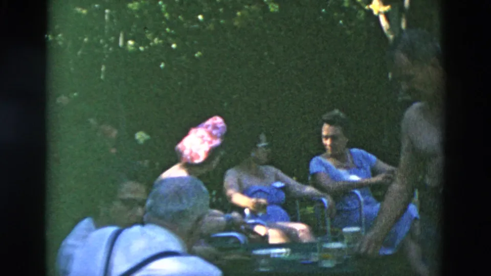 Outdoor 8mm frame of adults in lawn chairs under trees, with a standing shirtless figure cropped at the right edge.