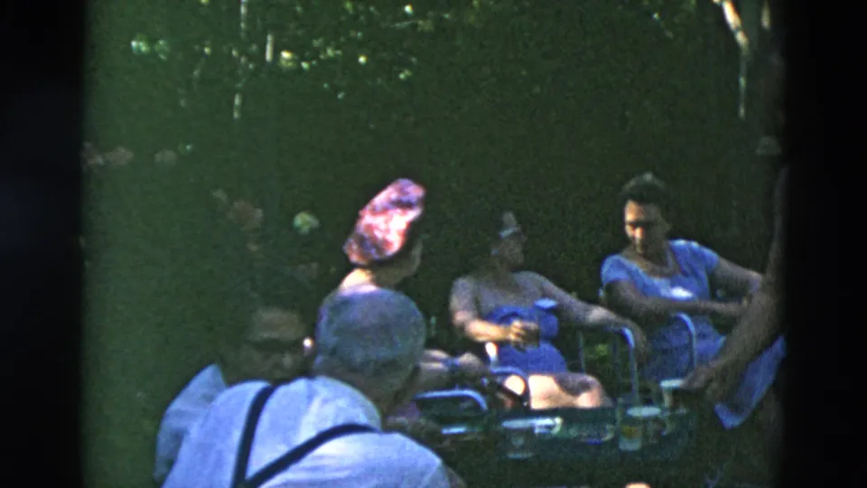 Adults in summer clothing sit near a table in a shaded backyard setting, with foliage and lawn chairs visible in 8mm home-movie footage.