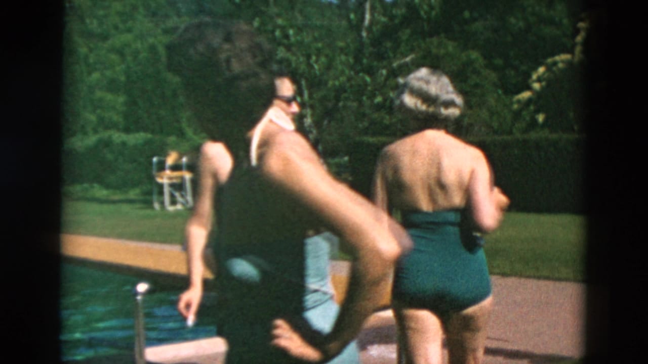 Adults in one-piece swimsuits and swim trunks move beside a backyard pool, with lawn and hedges in the background.