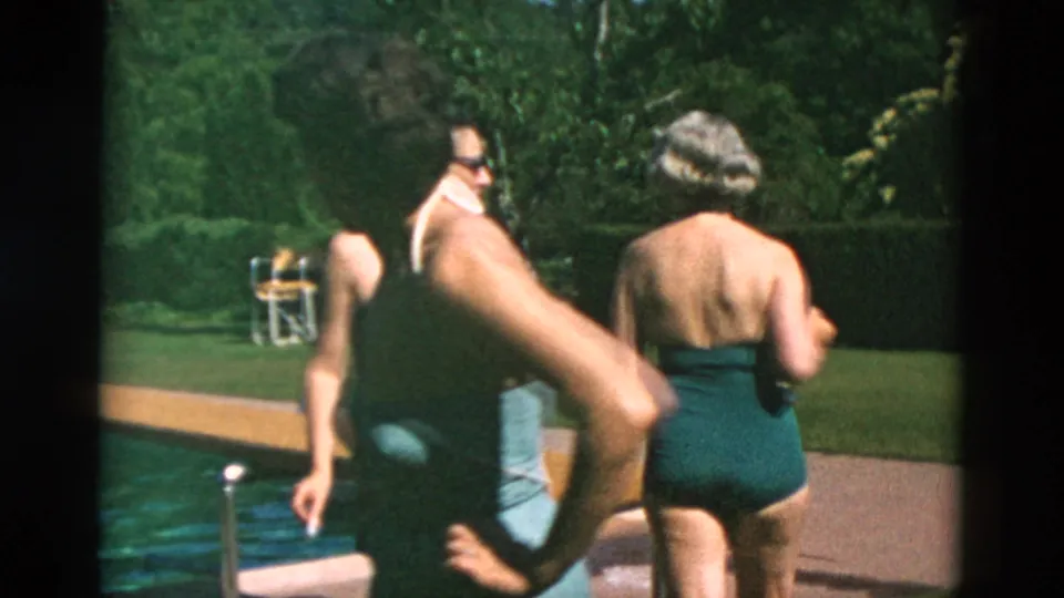 Adults in one-piece swimsuits and swim trunks move beside a backyard pool, with lawn and hedges in the background.