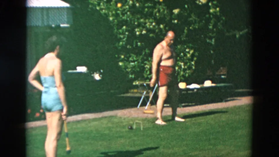 Two adults in swimwear stand on a sunlit lawn with croquet mallets, with poolside chairs, hedges, and garden foliage behind them.