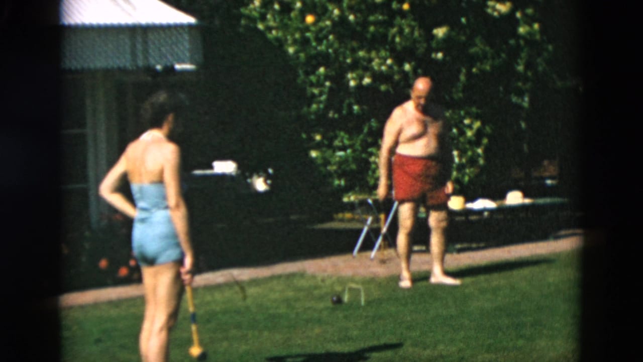 Woman in a blue swimsuit faces a shirtless man in red trunks on a lawn with croquet equipment.