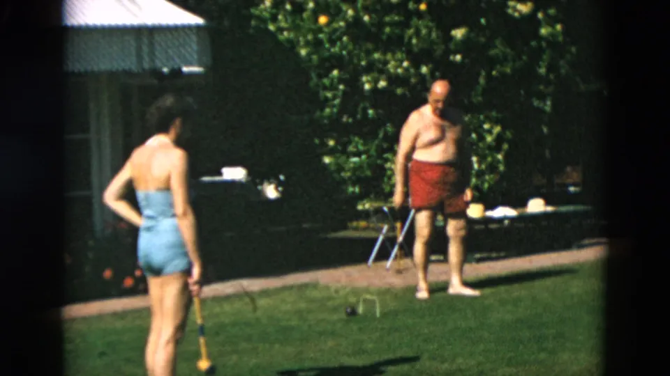 1950s 8mm home-movie frame of adults on a green lawn, with a woman holding a mallet and a shirtless man near croquet equipment.