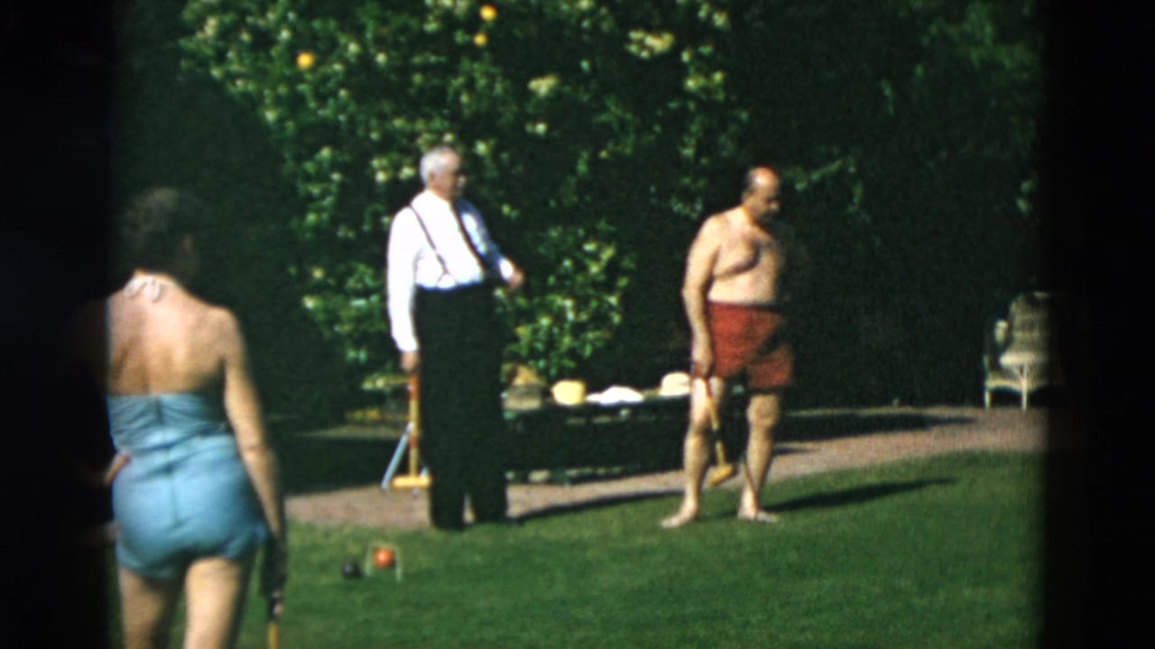 1950s 8mm frame showing a woman in blue swimwear, an older man in suspenders, and a shirtless man in red trunks on a garden lawn.
