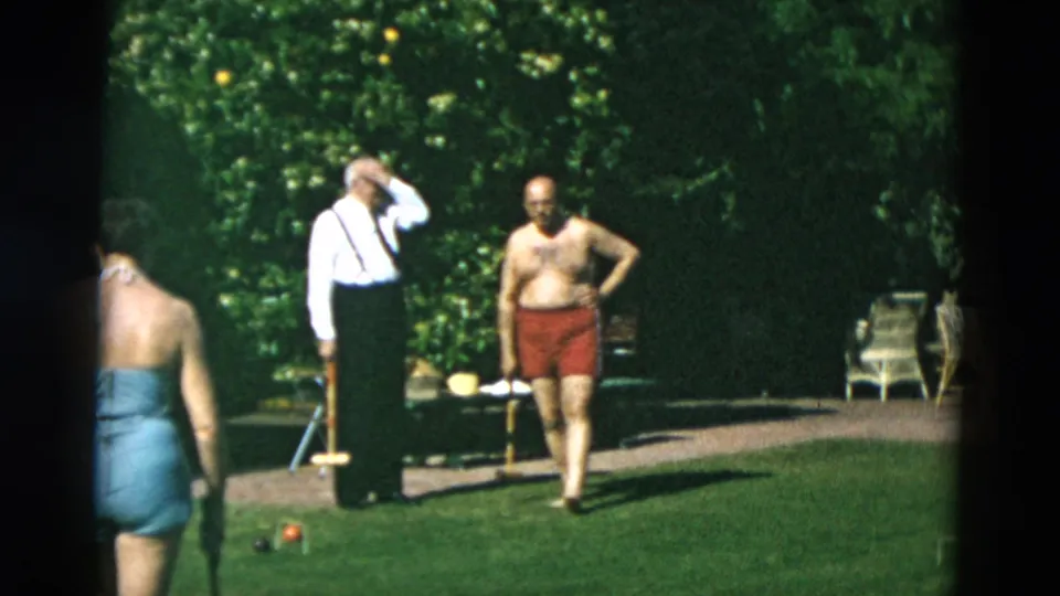 Home-movie frame of adults pausing on a croquet lawn, with one man in suspenders and another in red swim trunks near garden furniture.
