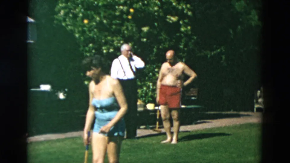 1950s home-movie frame with a woman carrying a croquet mallet in front of two men standing on a sunny garden lawn.