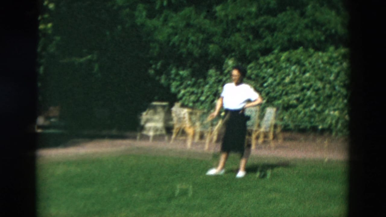 Woman standing on a green lawn with croquet equipment and patio chairs behind her.