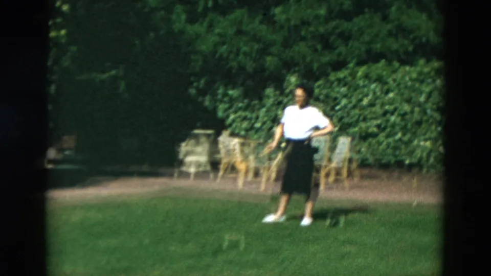 Woman in a white top and dark skirt stands on a sunlit lawn near croquet equipment and outdoor chairs in archival home-movie footage.