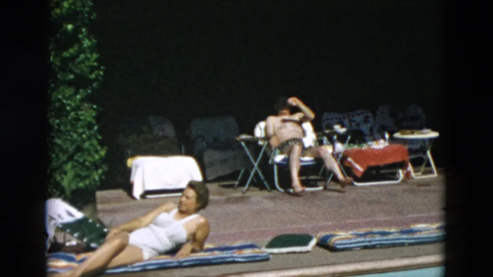 Adults lounge by a poolside area with towels, folding chairs, and small tables in sunlit 8mm home-movie footage.