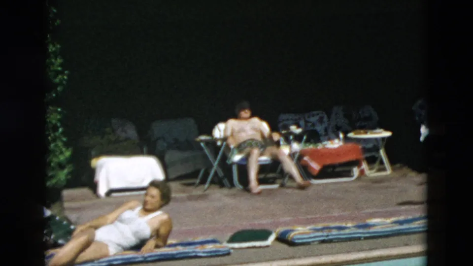Adults sunbathe around a poolside patio with striped towels, folding furniture, and shaded seating in archival home-movie footage.