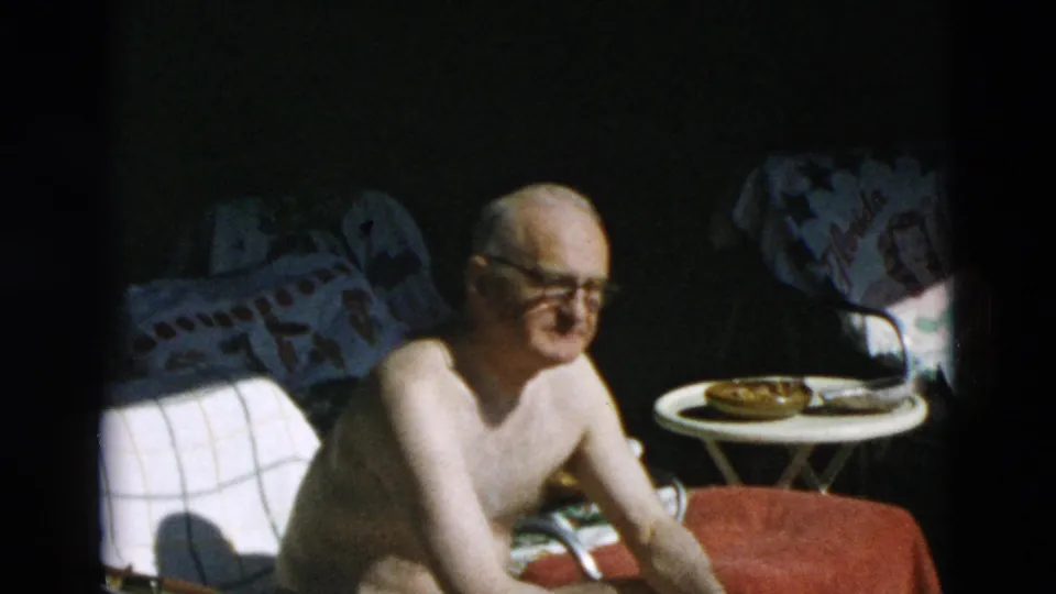 An older man wearing glasses smiles near poolside chairs, a small table, and towels in soft 8mm home-movie footage.